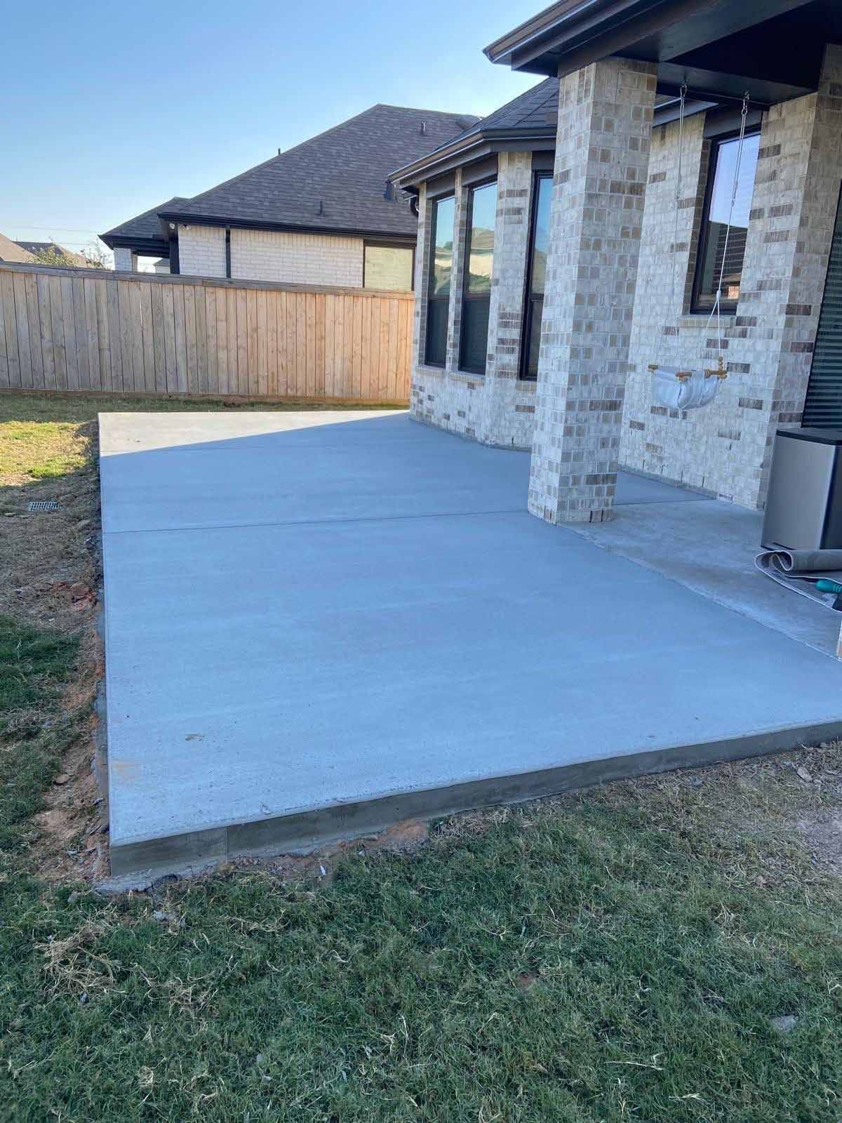 A gray concrete patio outside a house with a brick column and a wooden fence in the background.