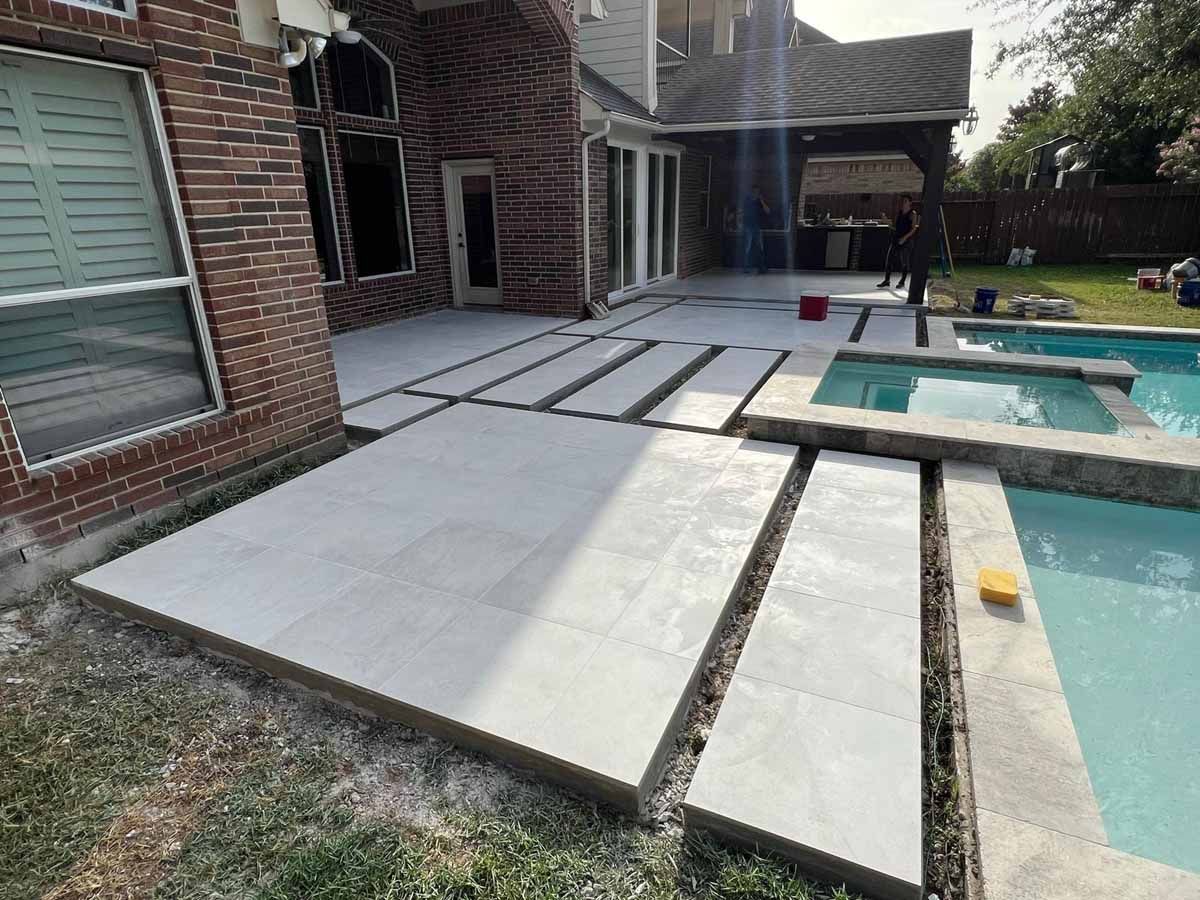 Patio with concrete pavers next to a pool and house with brick and glass windows.