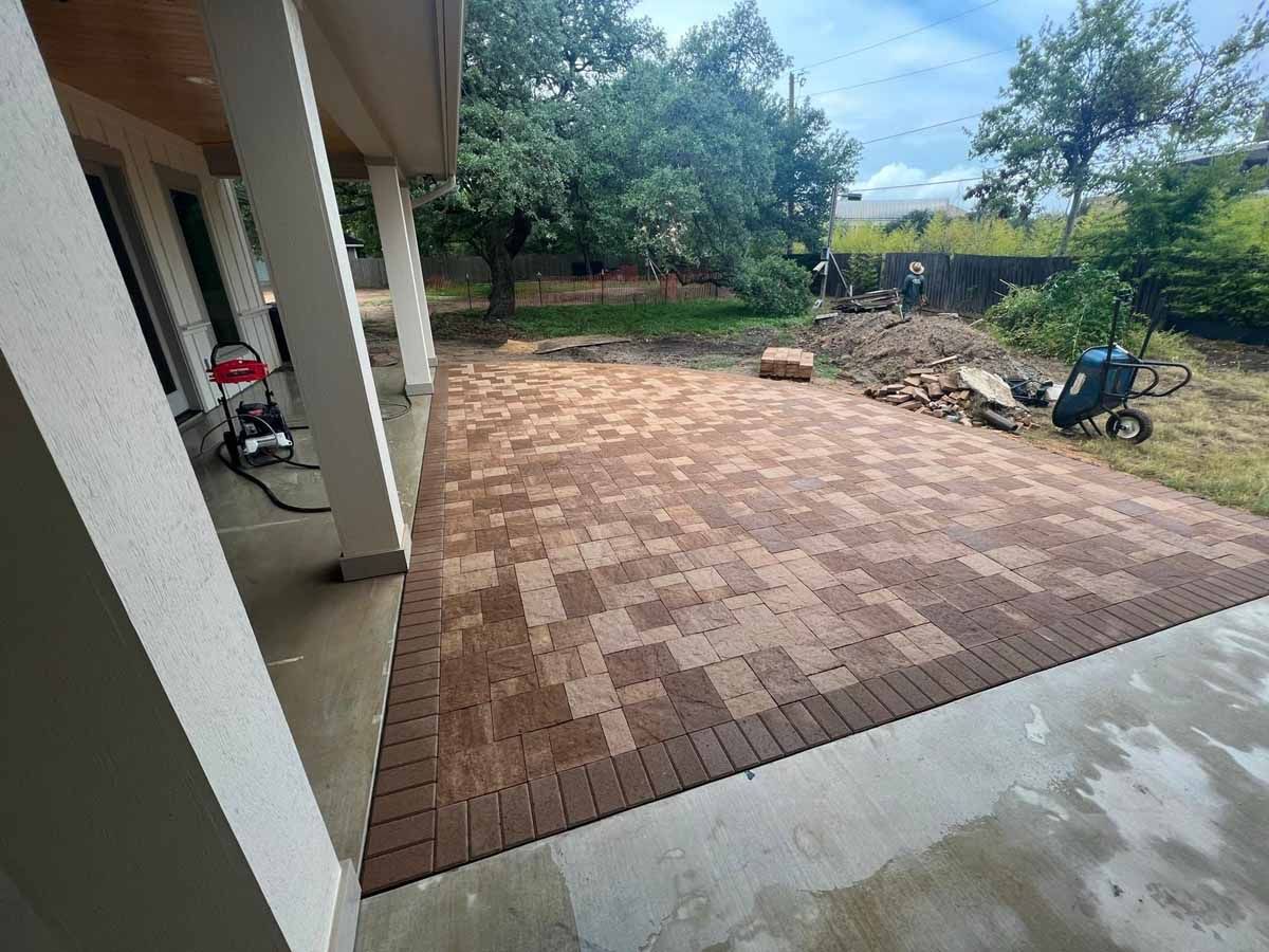 Brick patio under a porch, surrounded by yard and trees; various construction materials visible.