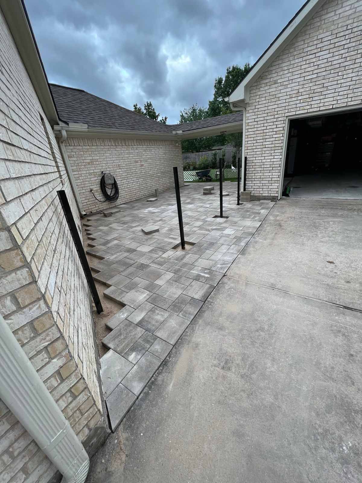 Paver patio next to a brick house and garage with black posts installed, and a cloudy sky.
