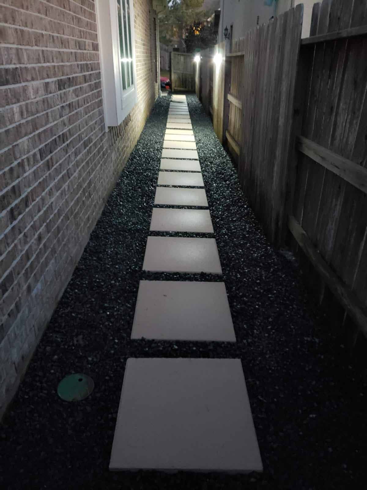 A lit pathway between a brick wall and wooden fence, with stepping stones on black gravel.