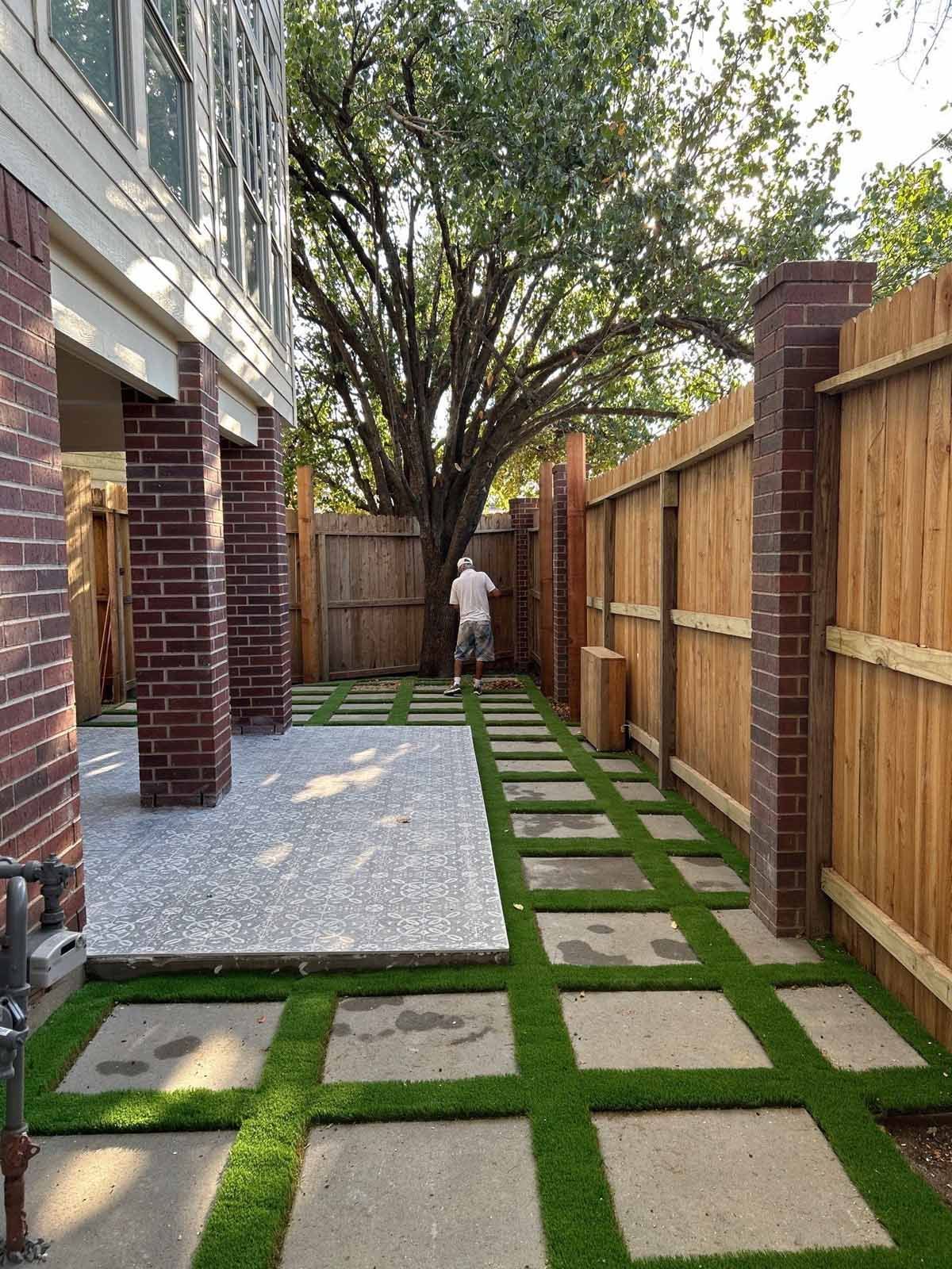 Backyard with artificial grass stepping stones and a wooden fence, person in the distance near a large tree.