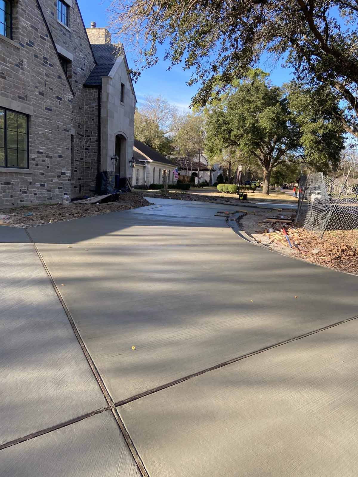 Newly poured concrete driveway leading to a house, with trees and other houses visible in the background.