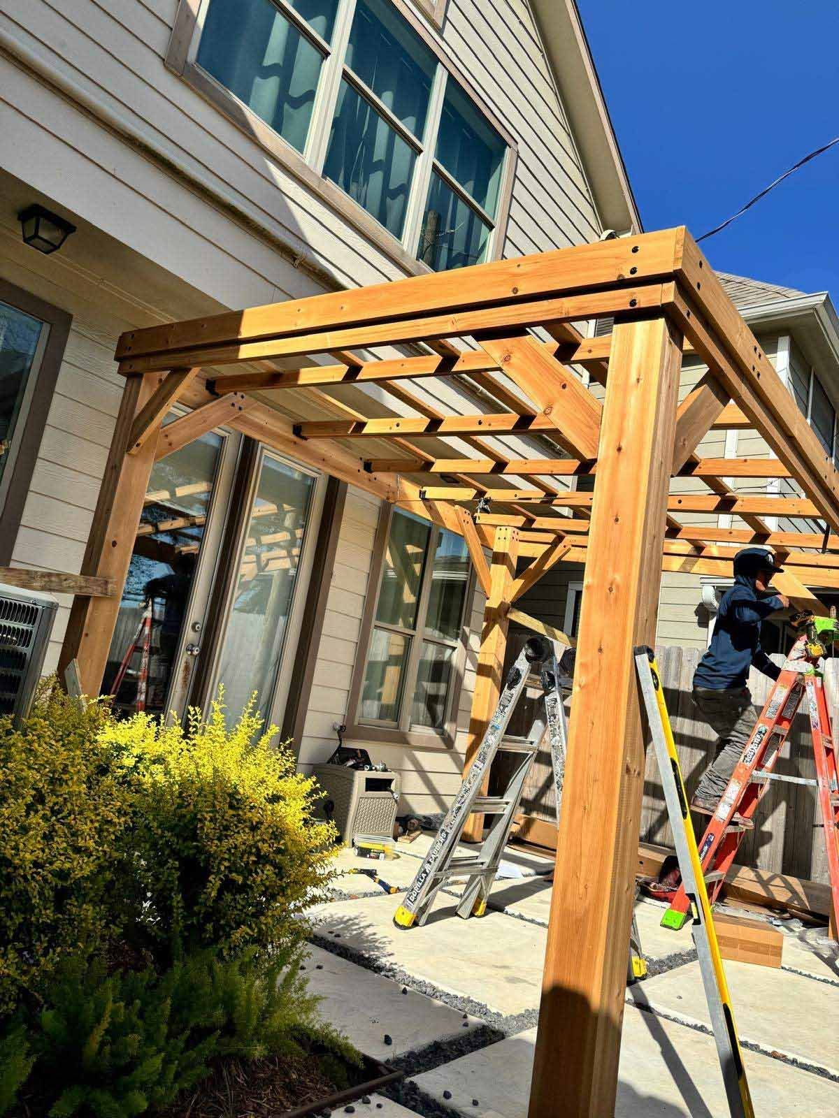 Wooden pergola being built outdoors, near a house with windows. Two ladders and a person working on the structure.