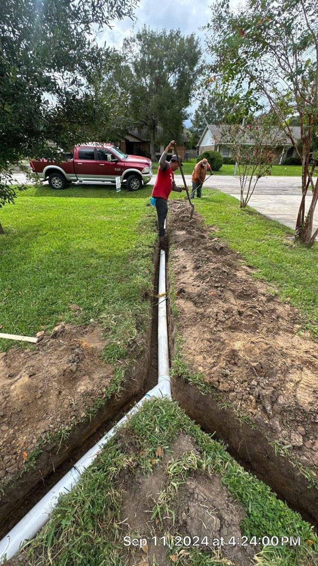 Workers installing white pipes in trenches along a grassy yard, truck parked in background.