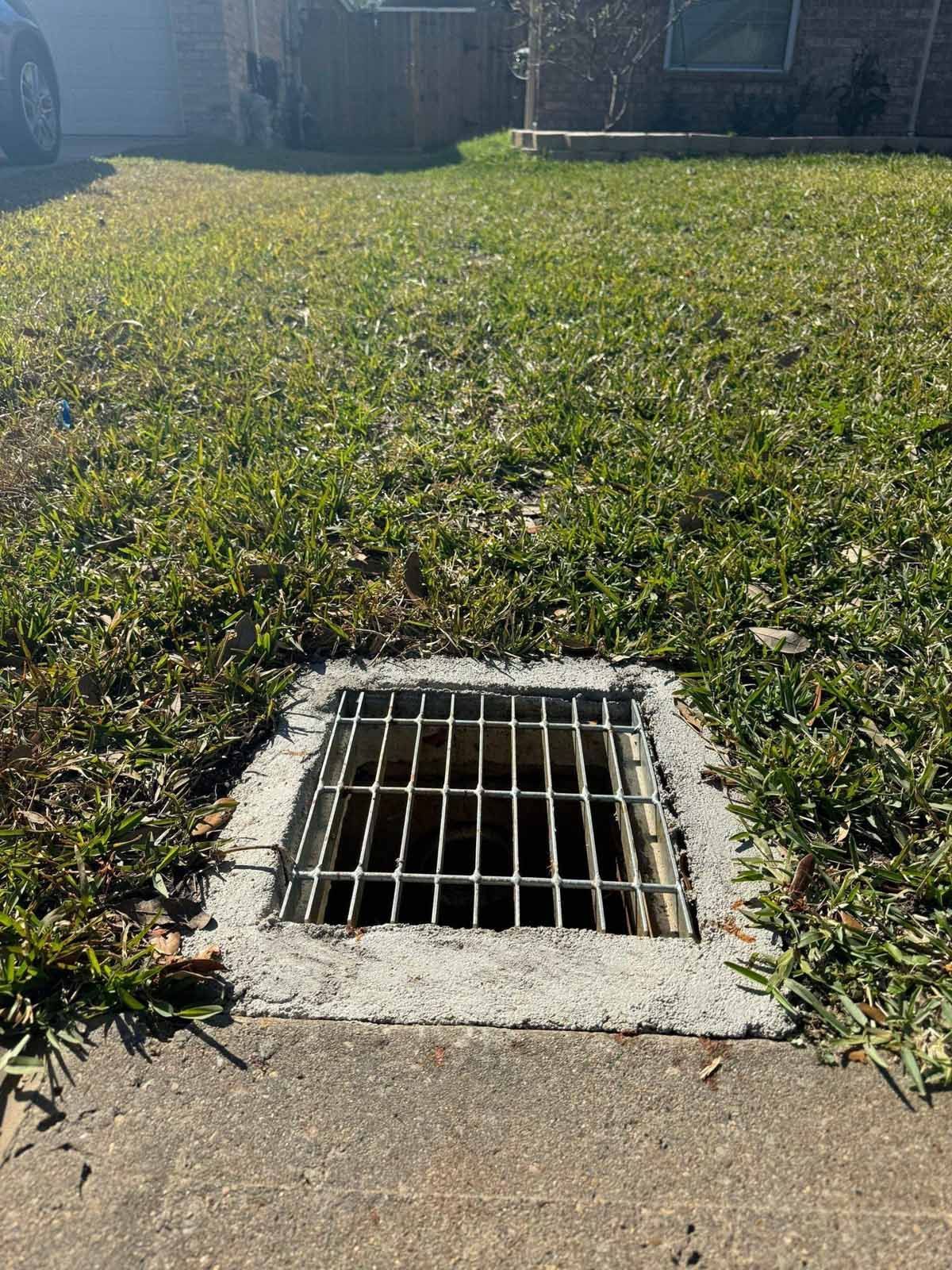 Square metal drain in a concrete frame set in grass. Sidewalk in foreground, house in background.