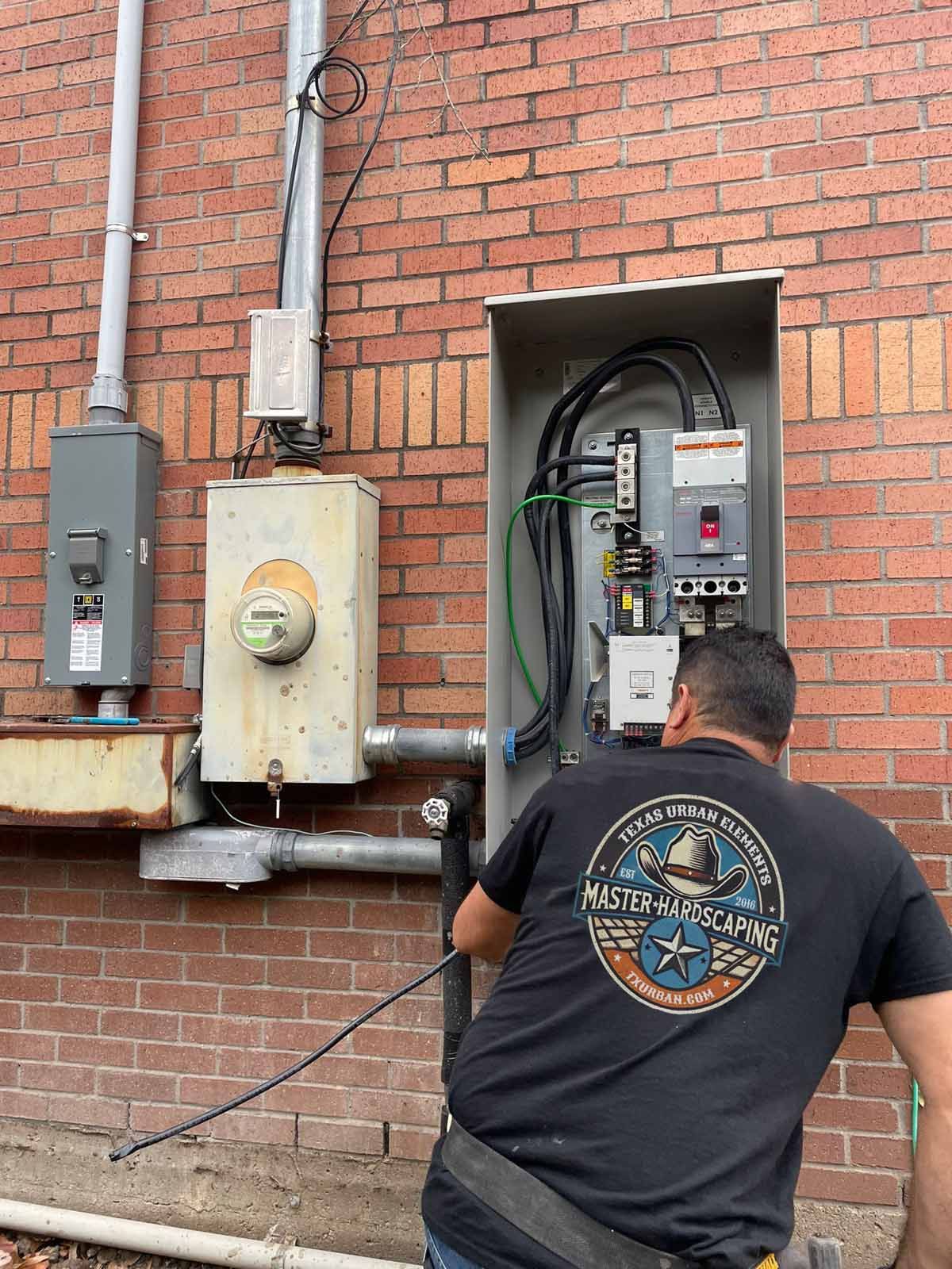 Electrician working on electrical panel mounted on brick wall.