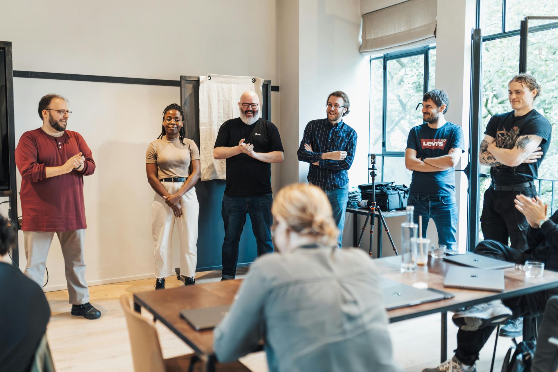 Small group standing in a bright meeting room, listening during a discussion around a table.