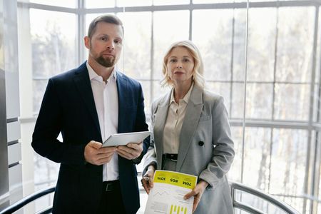 Two professionals standing in a bright office lobby, holding documents and looking forward.