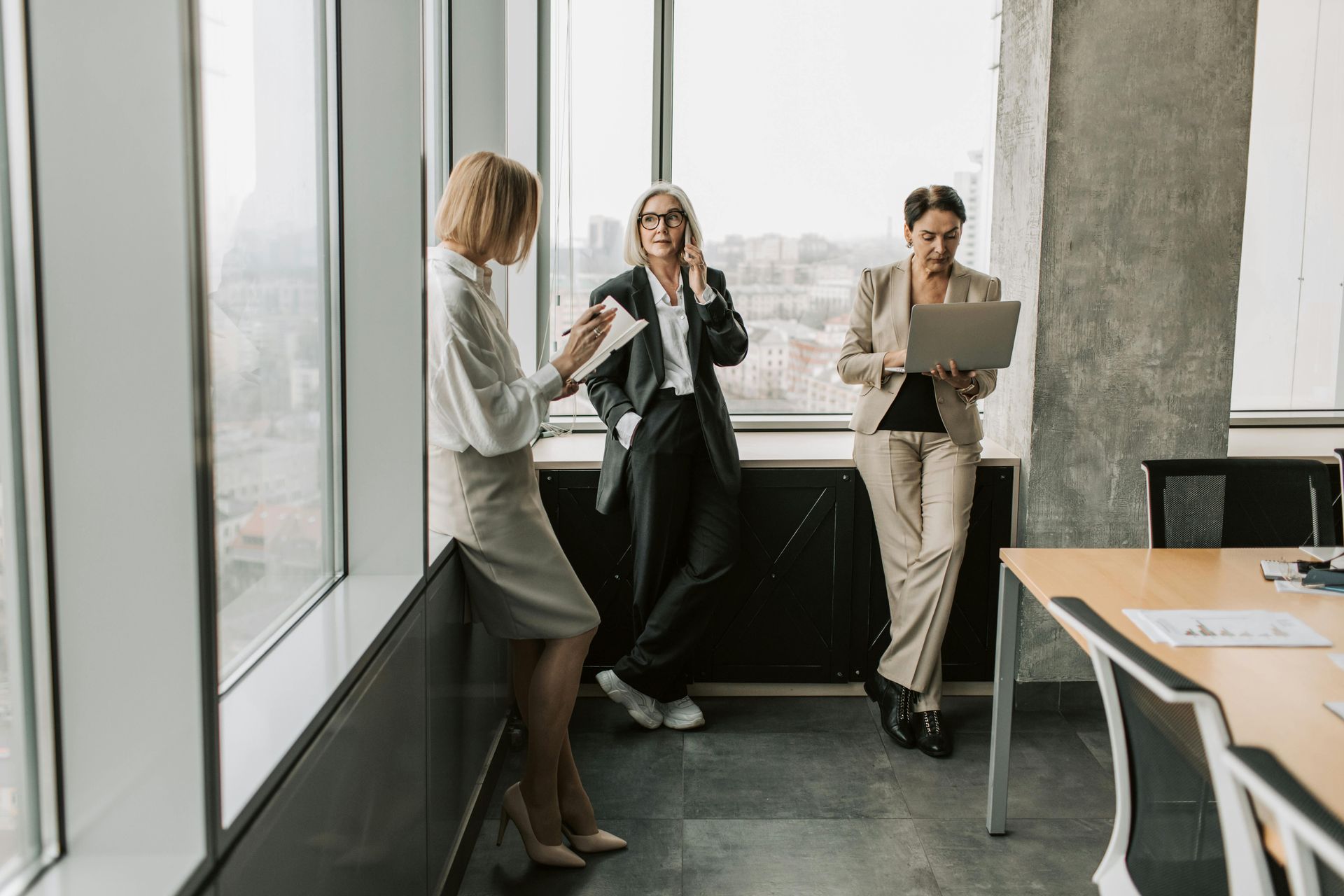 Three colleagues talking in a bright office near a window, one holding a laptop.