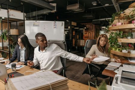 Three coworkers in an office, reviewing papers at a shared desk with shelves and equipment nearby.