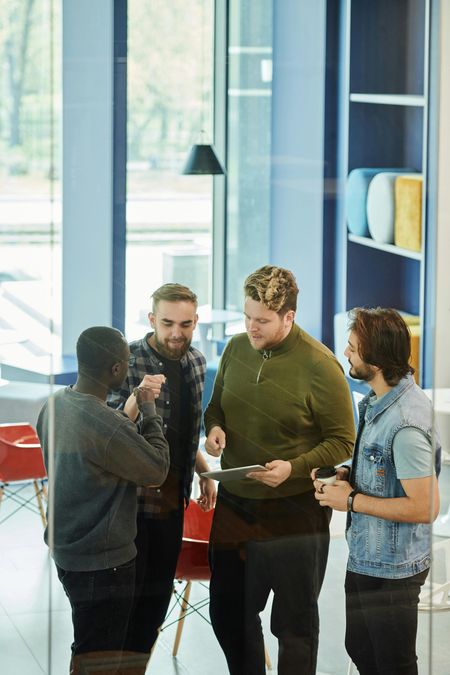 Four coworkers discuss notes in a bright office lobby beside red chairs and blue shelving.