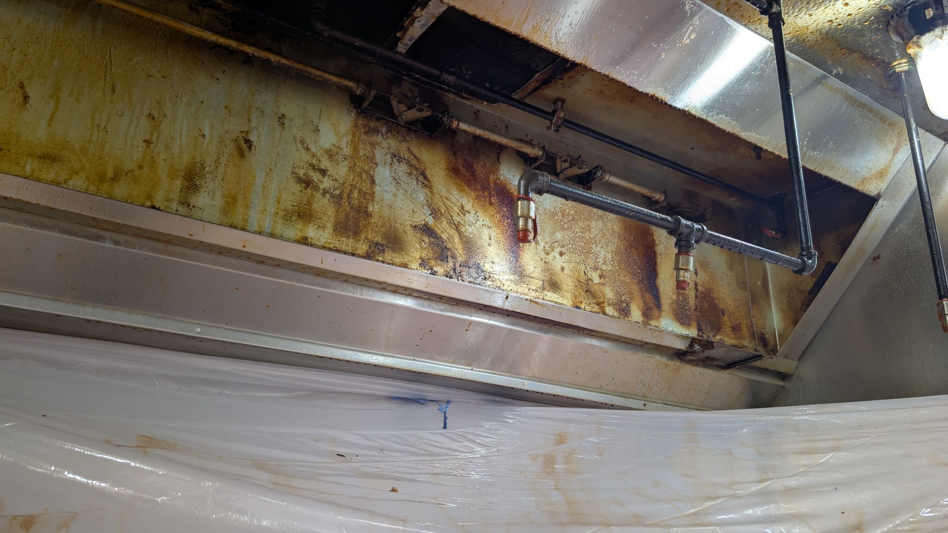 Person cleaning a commercial kitchen exhaust hood system, which is draped in plastic sheeting to contain cleaning debris.