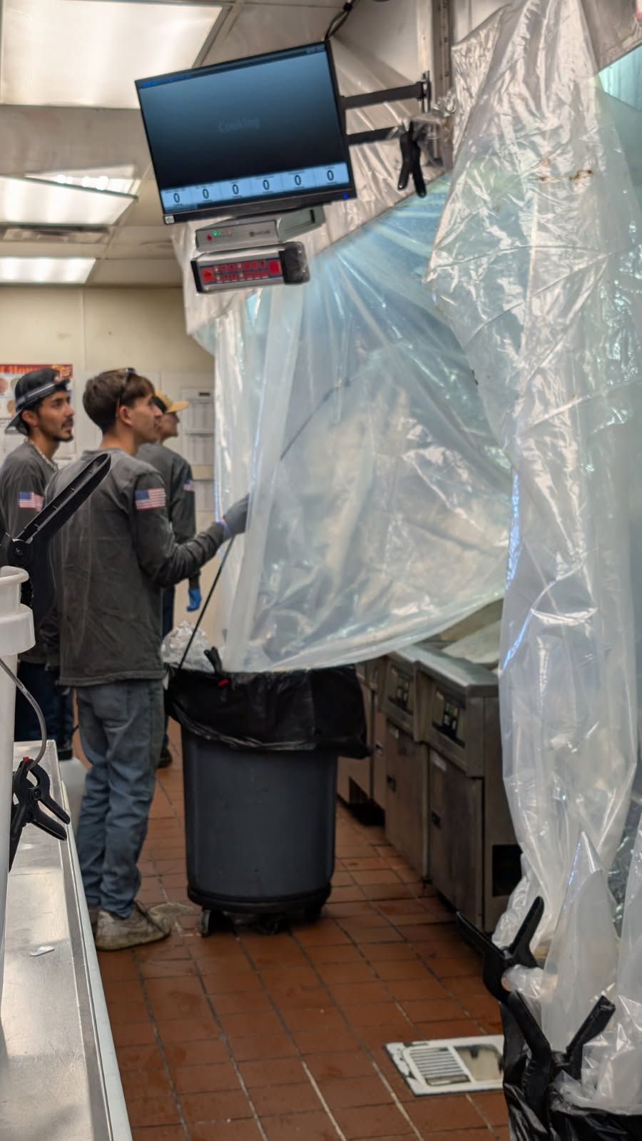 Three people in a commercial kitchen, one using plastic sheeting to seal a work area. The setting is brightly lit.