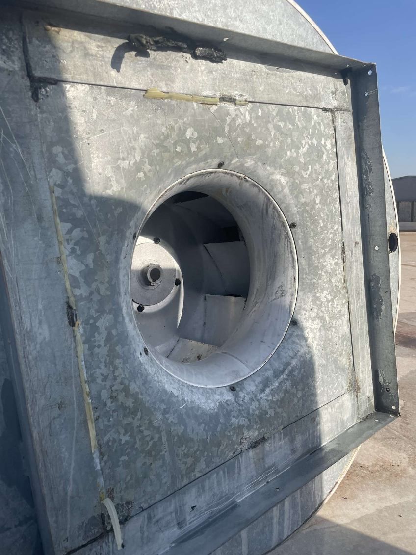 Close-up view of a galvanized metal centrifugal fan. The circular fan blades are visible within a large, square housing.