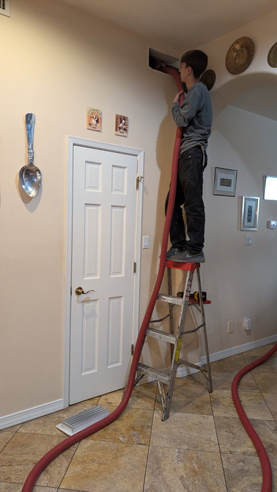 A person on a stepladder is working in a wall vent, holding a red hose. They are in a brightly lit room, next to a white door.