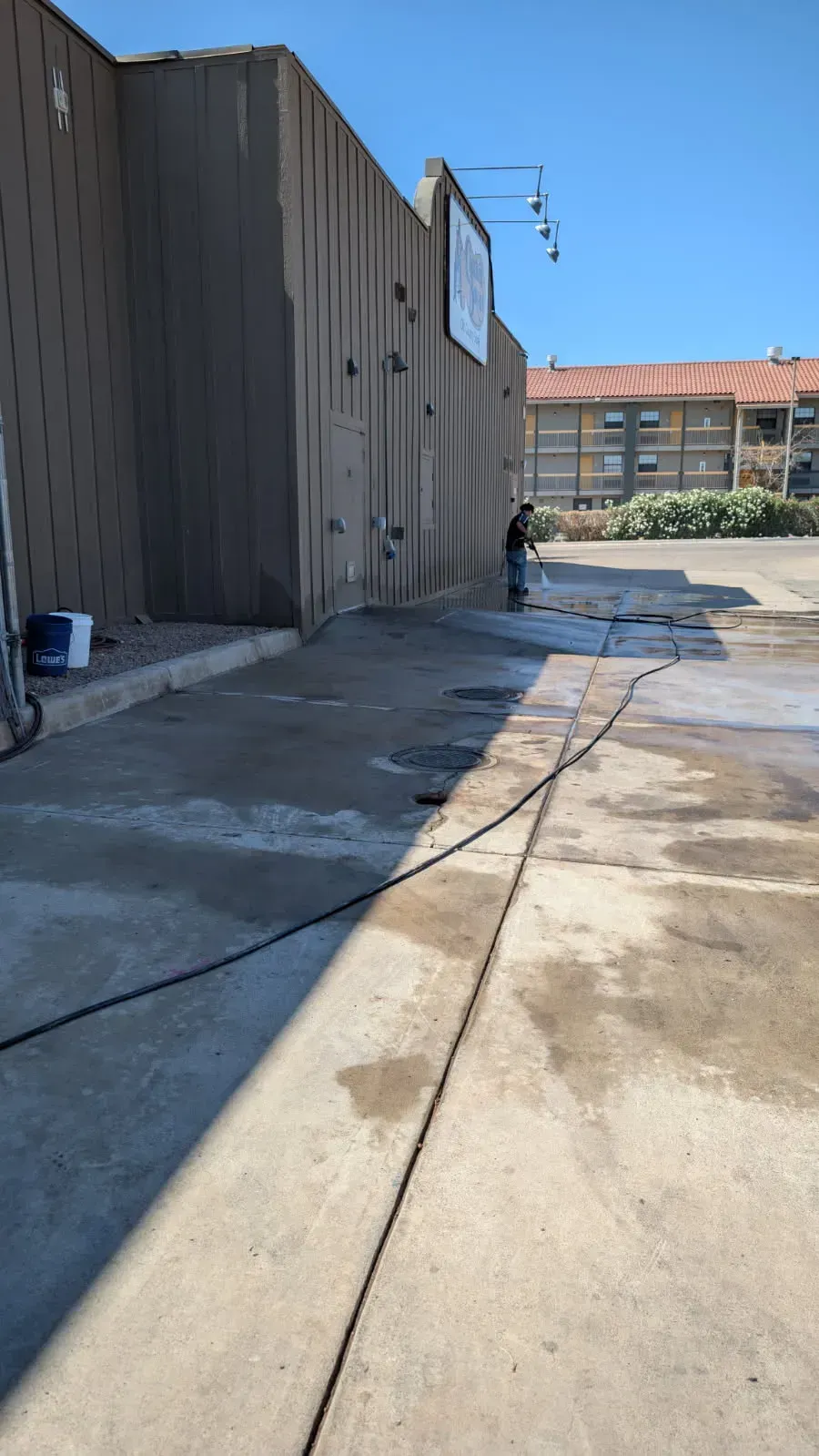 Person washing a concrete wall with a hose outside. Building is gray, sky is blue.