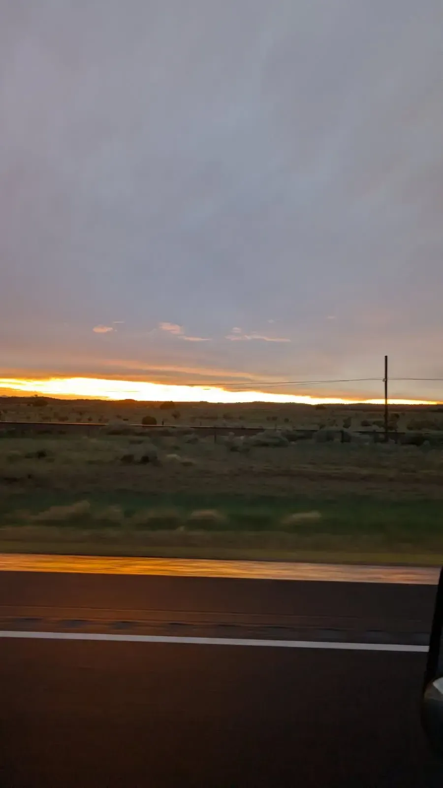 Sunset over a grassy plain, seen from a car. Orange glow beneath a cloudy gray sky.