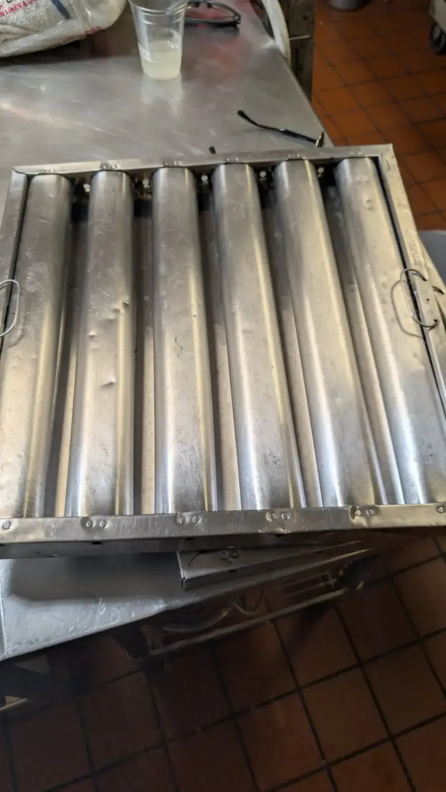 A stainless steel kitchen exhaust hood filter sits on a metal countertop. It is rectangular with multiple vertical baffles.