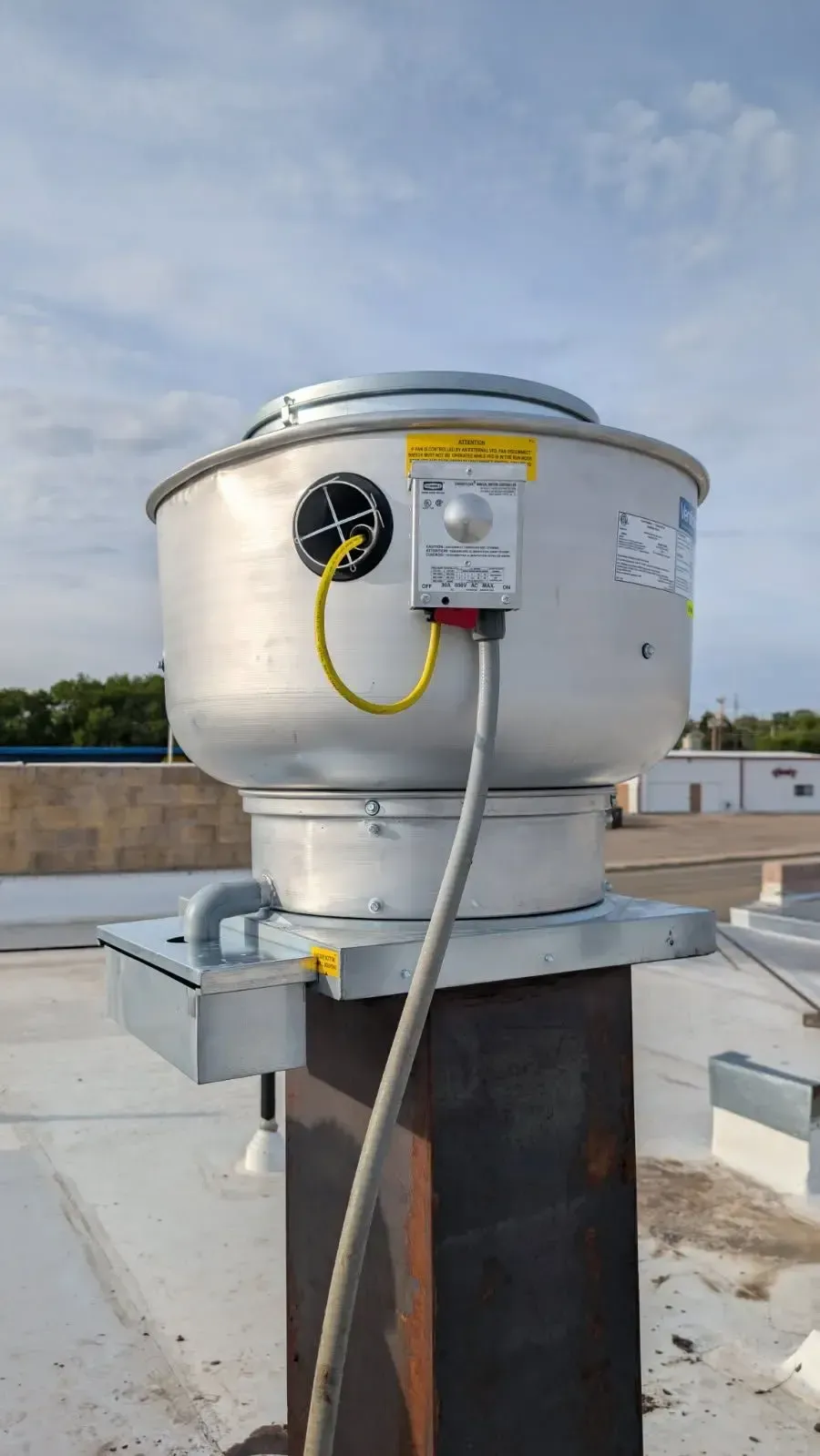Rooftop industrial exhaust fan with a metal housing and control panel. The fan sits atop a metal pillar on a flat roof.