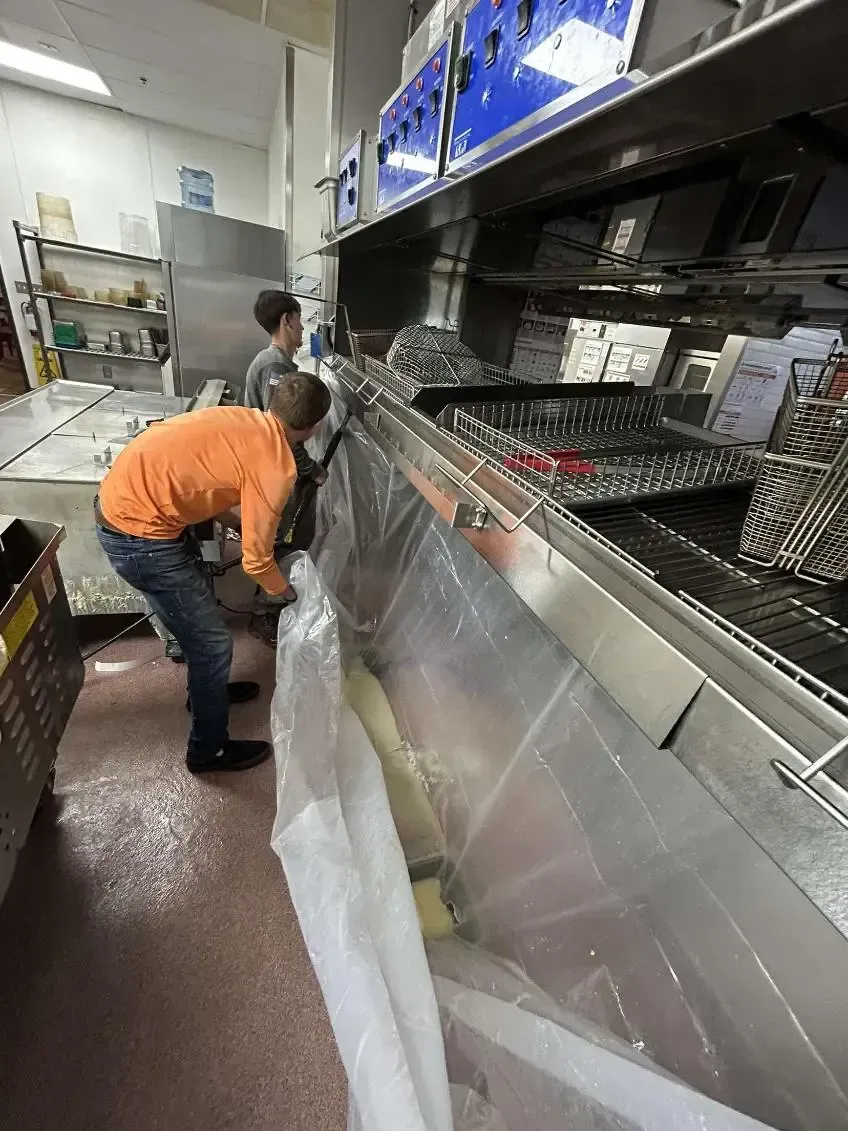 Two people cleaning a commercial kitchen, collecting debris in a clear plastic bag under a stainless steel hood.