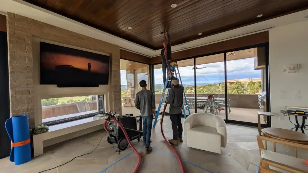 Two men set up equipment in a modern living room with large windows. A large screen displays an image above a fireplace.
