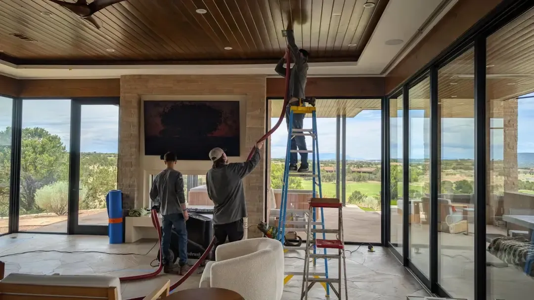 Three workers cleaning a ceiling in a large room with floor-to-ceiling windows overlooking a scenic landscape.