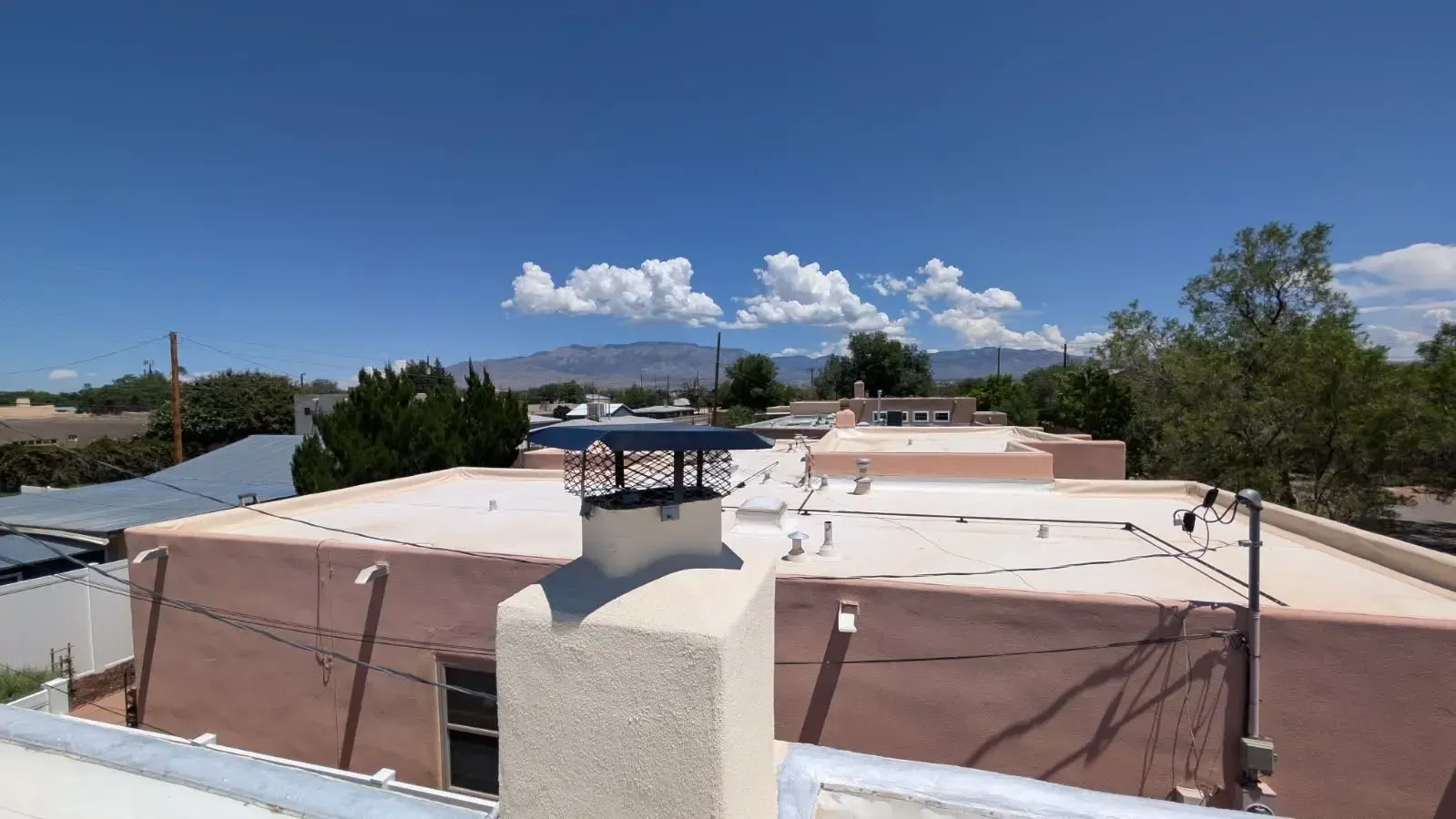 A view of a flat roof with a chimney under a blue sky with scattered clouds. Distant mountains are visible.