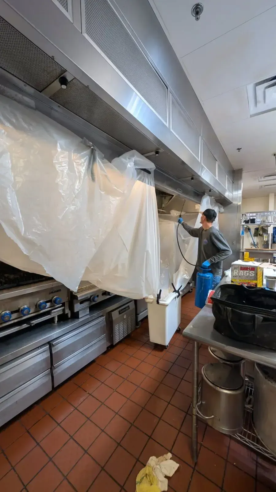 A person sprays a kitchen hood wrapped in plastic in a commercial kitchen. They wear blue protective clothing.