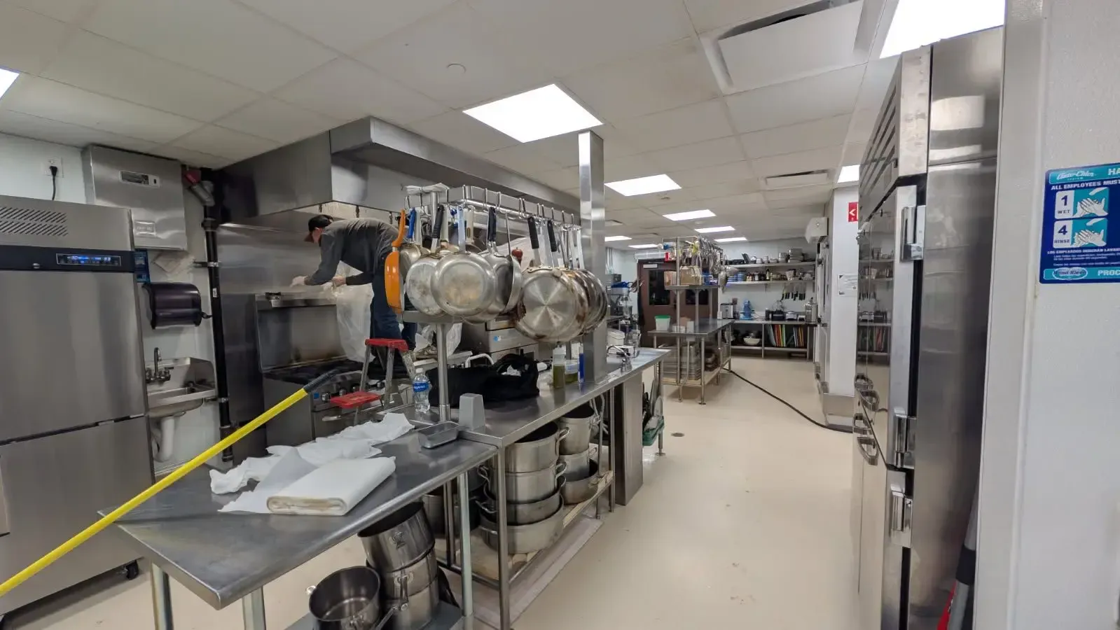 A commercial kitchen with stainless steel appliances and hanging pots. Long, narrow space with white walls and bright lights.