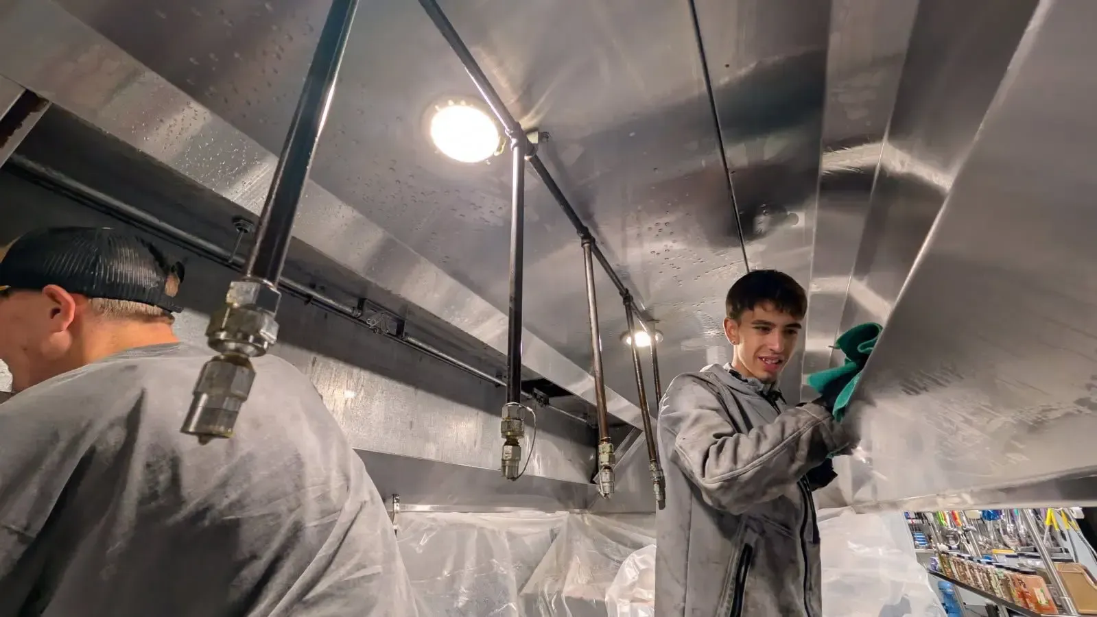 Two people in grey overalls working inside a metal structure, one cleaning the ceiling with a green cloth.