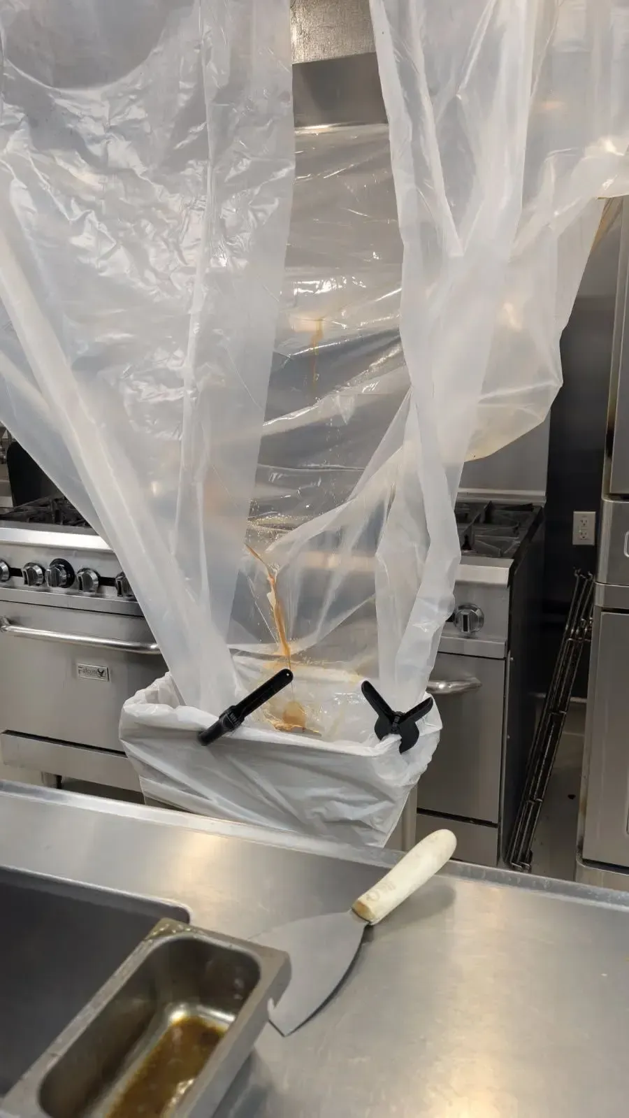 A commercial kitchen with plastic sheeting suspended over a stove and a metal bin held by black clips. A spatula sits on a stainless steel counter.