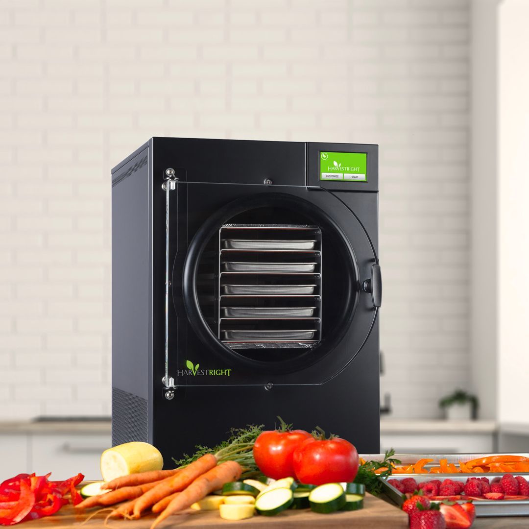 Black freeze dryer with a digital display, sitting on a kitchen counter with fresh vegetables in front of it.