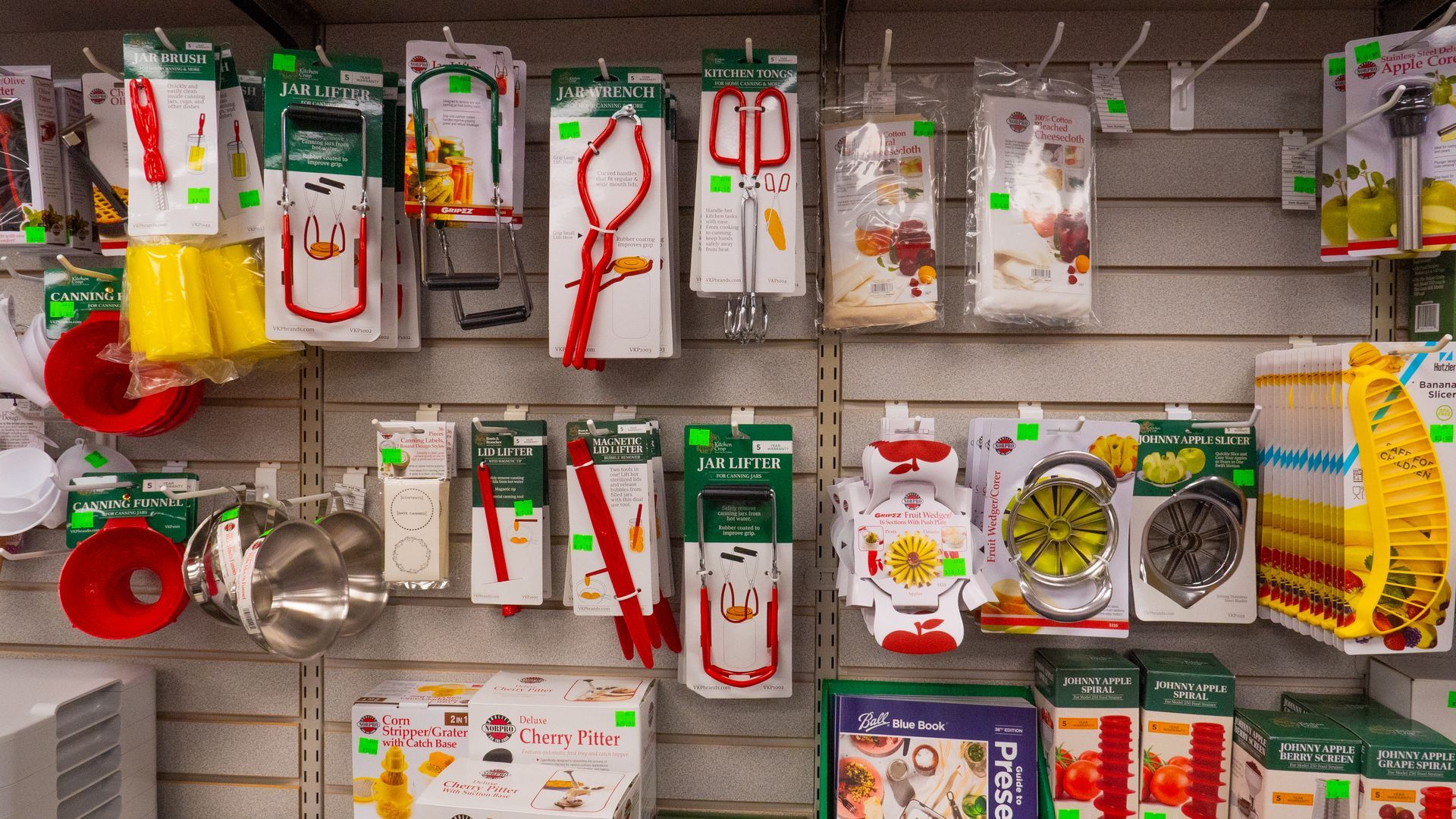 Shelves stocked with kitchen tools, including tongs, jar lifters, funnels, and citrus juicers, packaged with green and red labels in a store setting.