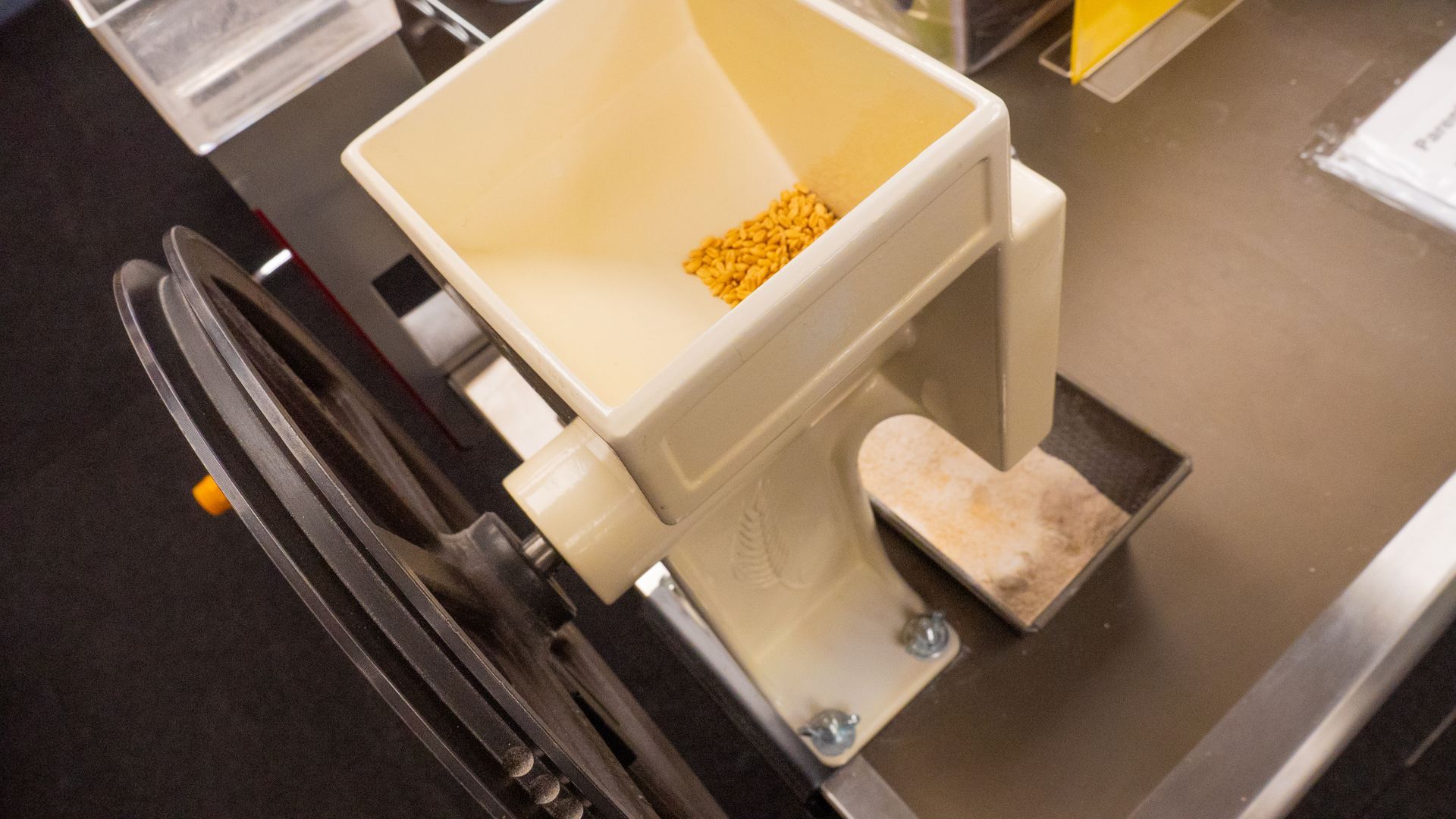 A close-up of a cream-colored grain mill with a hopper filled with grains. A hand crank wheel is on the left.