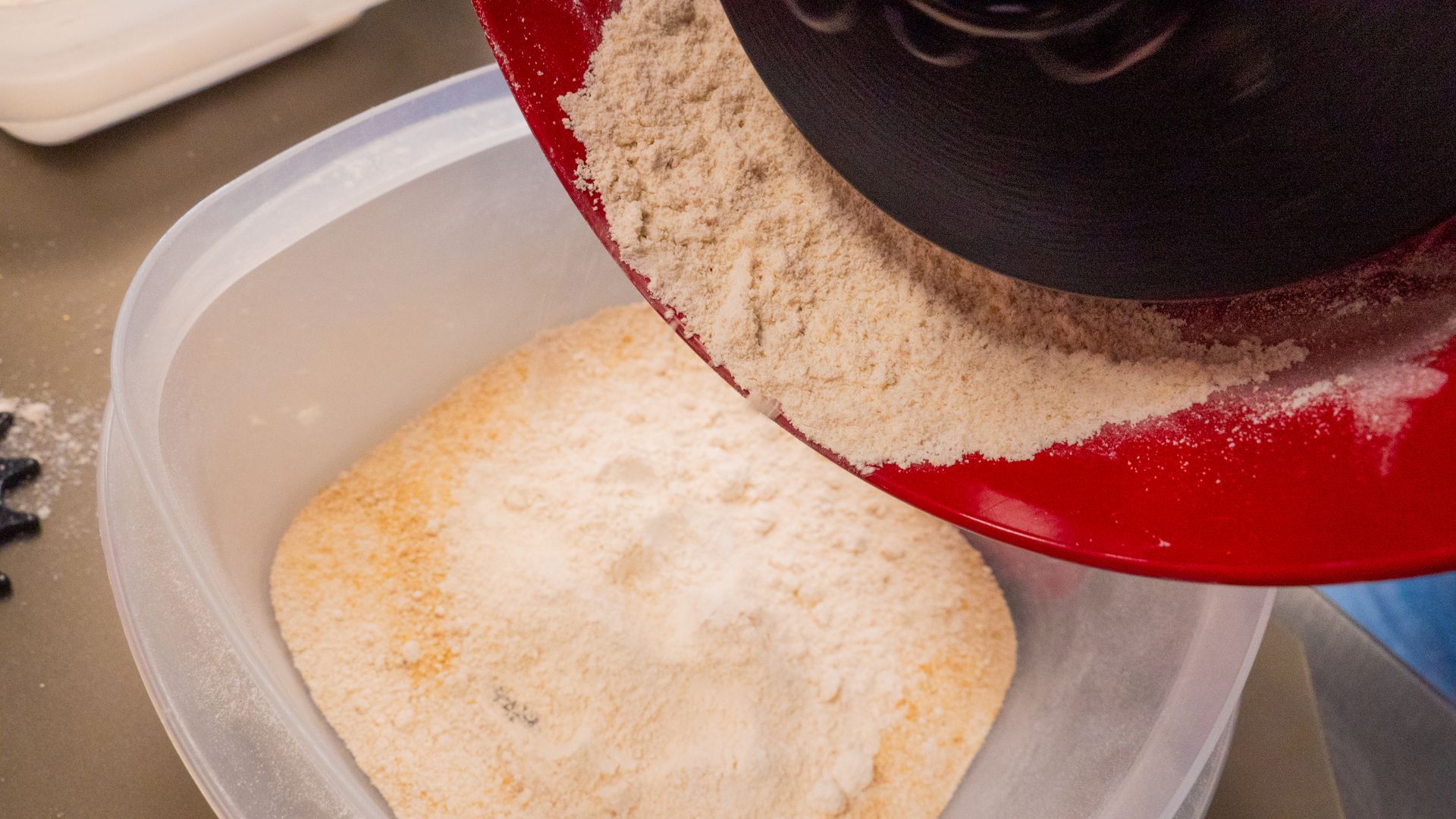 Red bowl pouring flour into a clear, rectangular container on a counter.