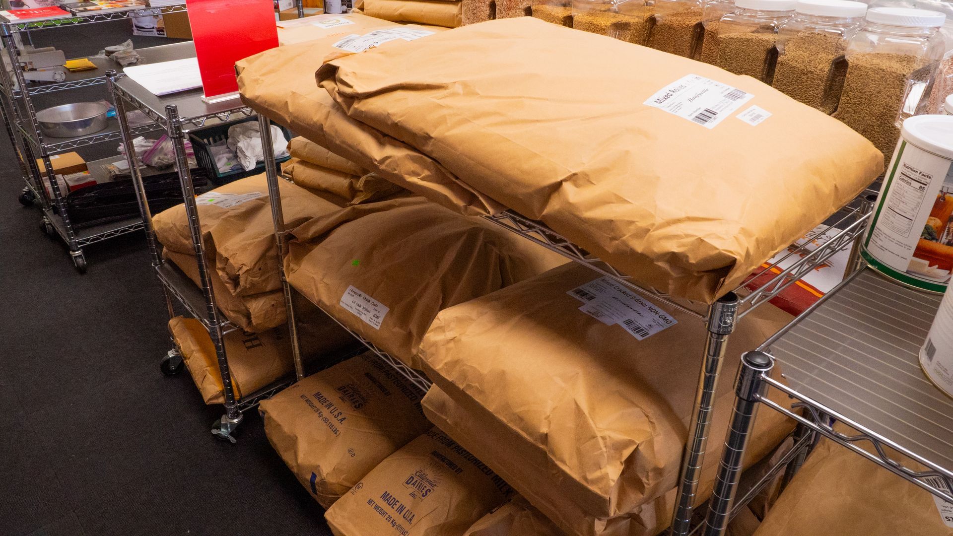 Bags of flour stacked on metal shelving in a commercial kitchen or storage area. Brown paper bags are labeled with text.
