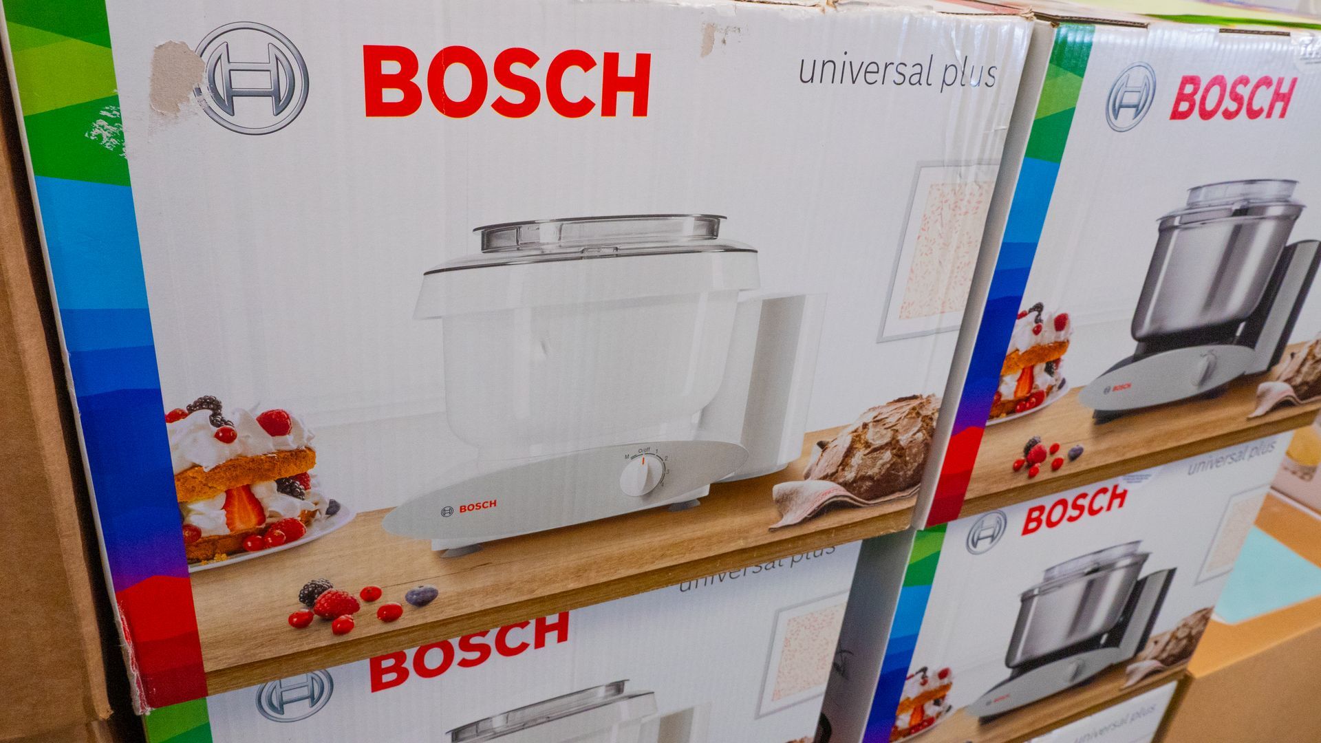 Boxes of Bosch Universal Plus mixers stacked on a shelf. The boxes are white with the appliance pictured and the brand name in red.