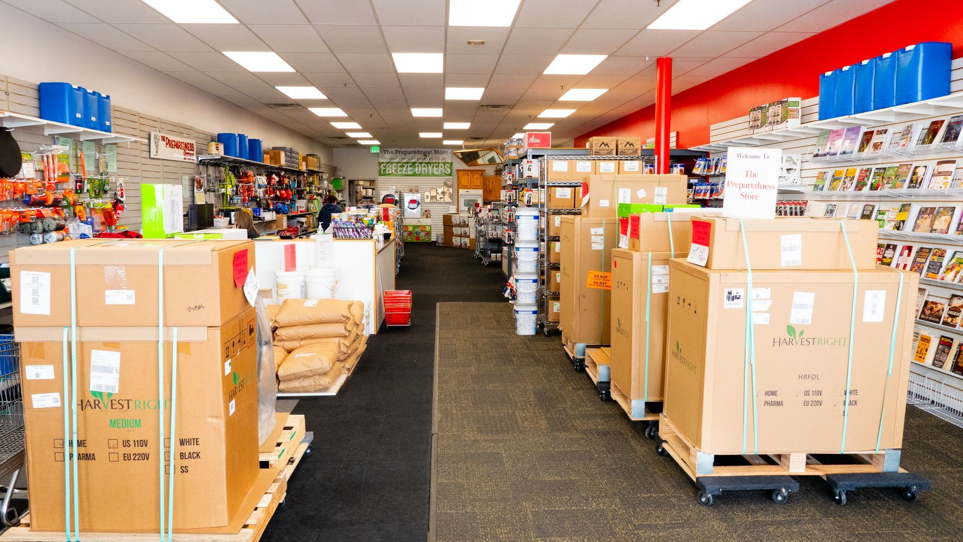 Inside a retail store, boxes on pallets line the aisles. Shelves hold various products; red and blue are prominent colors.