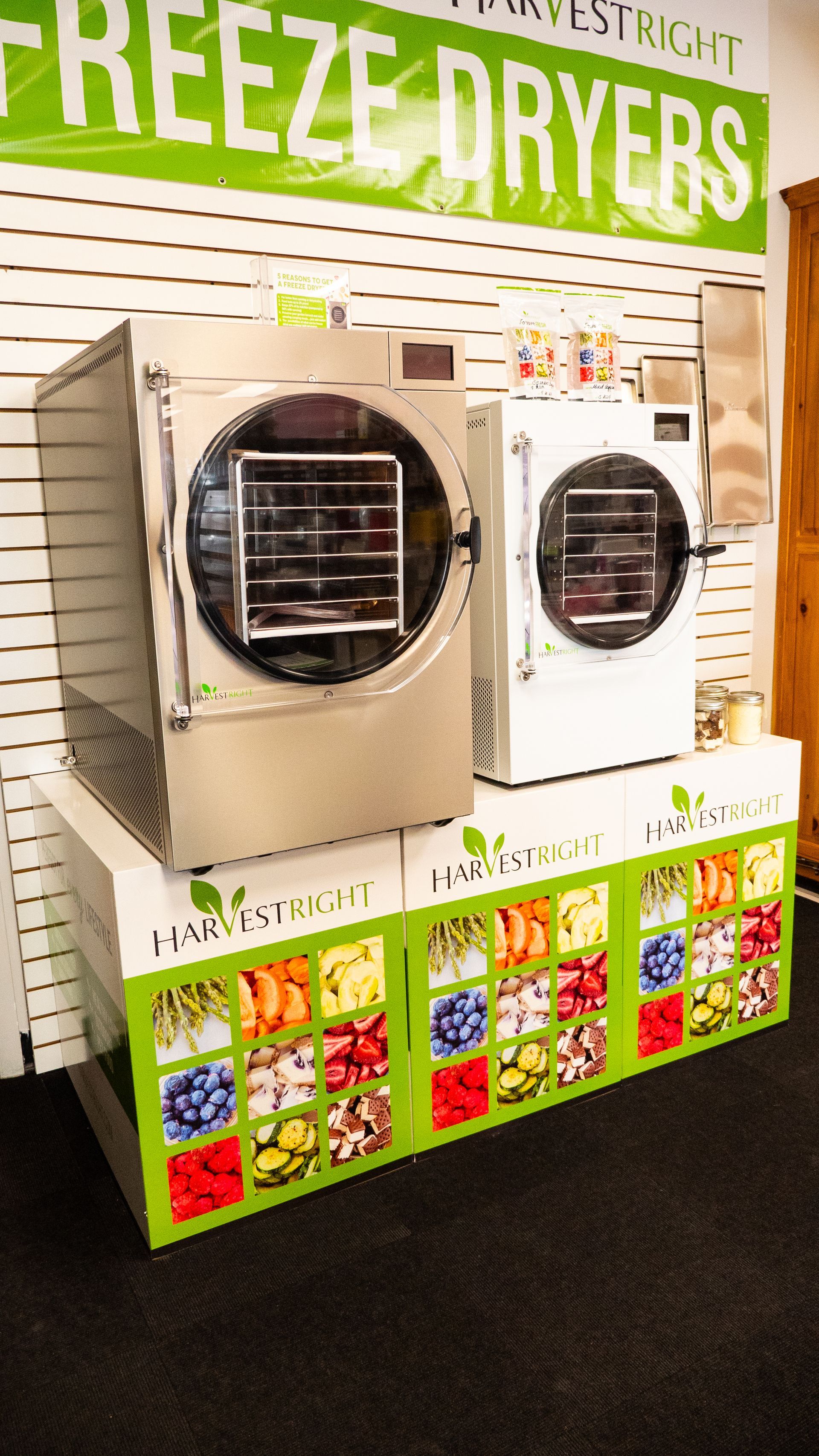Two freeze dryers, one silver and one white, sit atop a display with colorful fruit images. A sign reads 