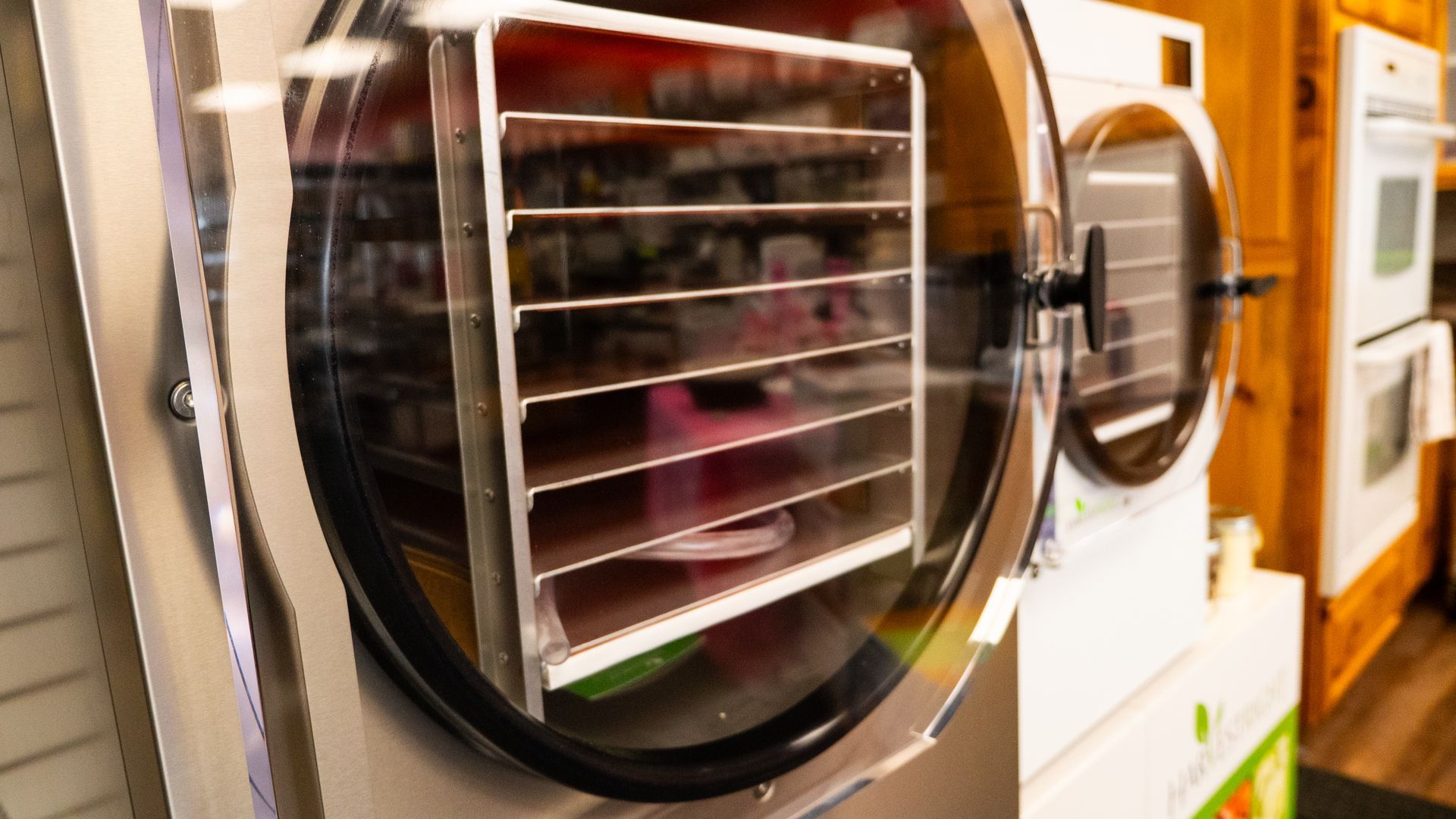 A freeze dryer with a clear door showing multiple trays inside. The machine is white and silver, set in a kitchen-like setting.