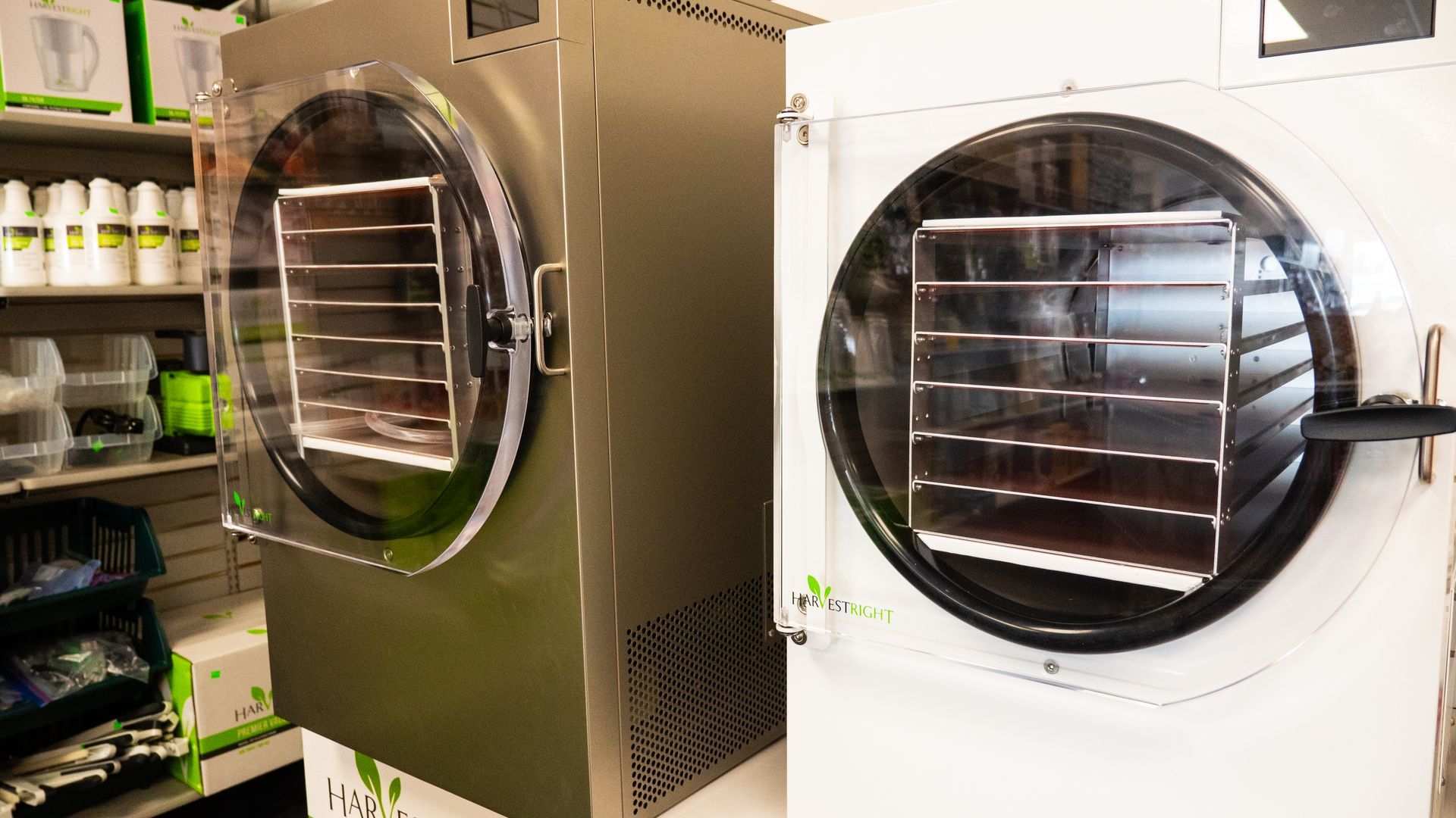Two silver and white freeze dryers with transparent doors, showing trays inside. They are in a store setting.
