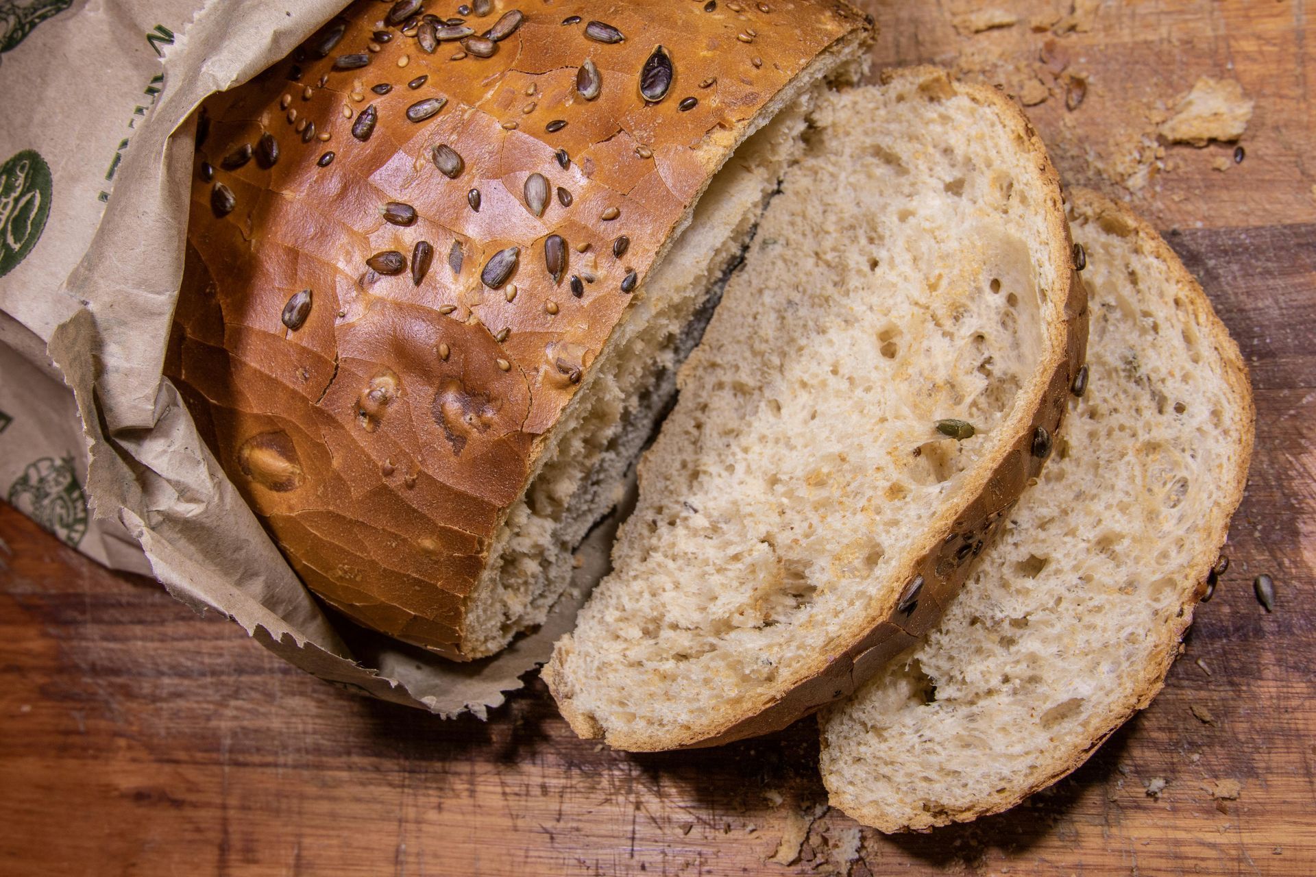 Loaf of seeded bread partially sliced on a wooden surface, inside a paper bag.