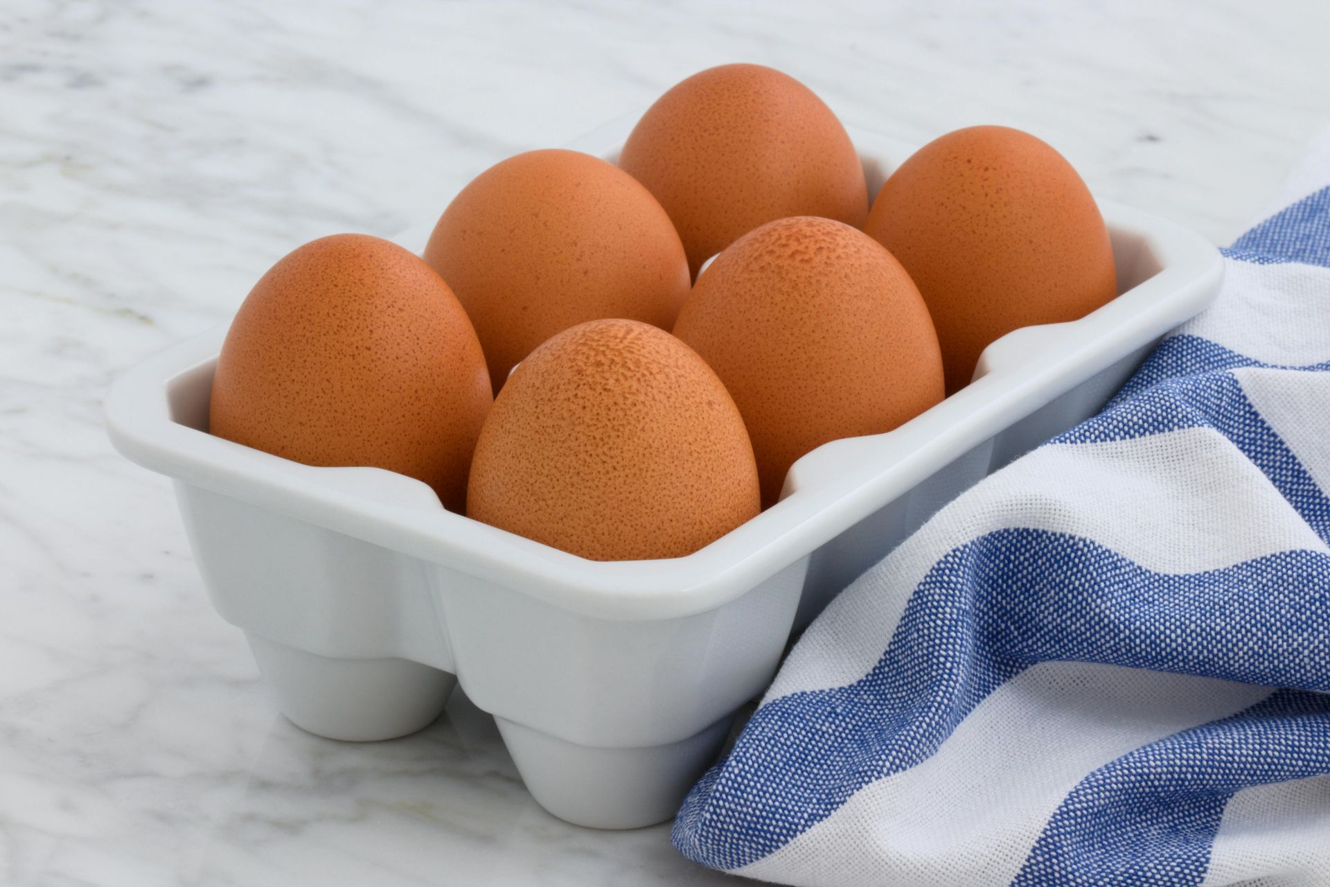 Six brown eggs in a white carton, next to a blue and white striped towel on a marble surface.