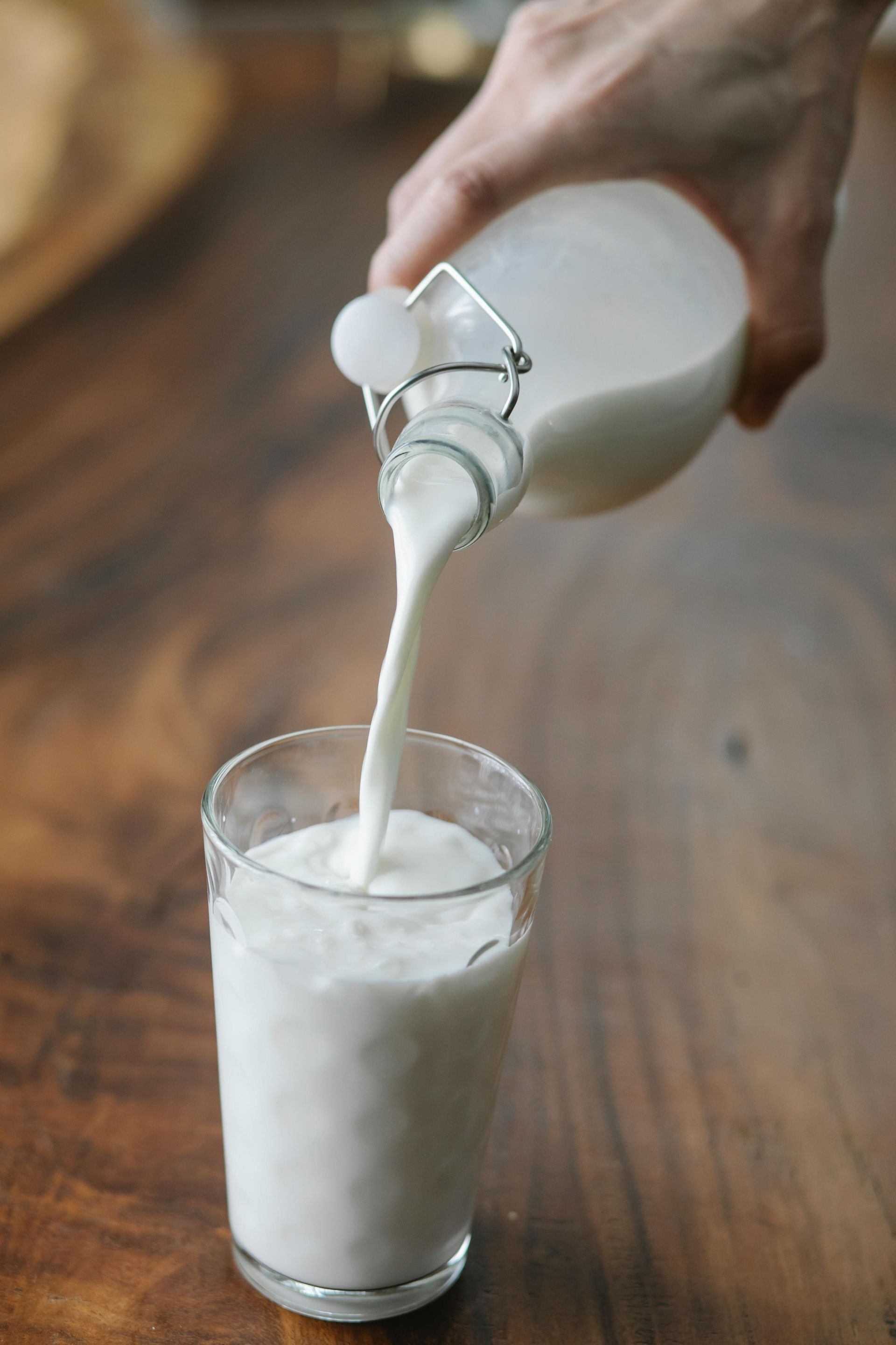 Person pouring white liquid, likely milk, from a bottle into a clear glass on a wooden table.