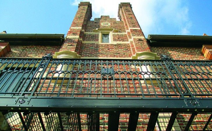 A low-angle view of a brick building with a black decorative metal balcony and two prominent chimneys against a blue sky.