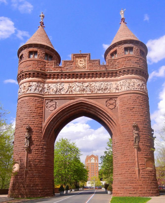 A large red sandstone Romanesque archway featuring twin towers, a frieze, and a tall brick building in the distance.