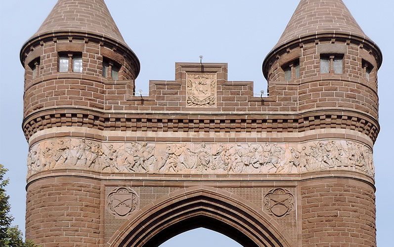 A tan, crenellated stone gateway with two round towers, featuring intricate carvings and calligraphy above a large arch.