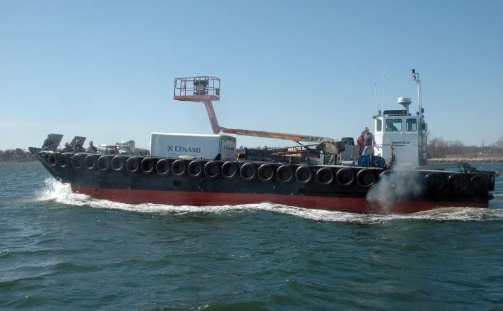 A work boat with black and red sides travels across open water, carrying a white container and a mounted crane arm.