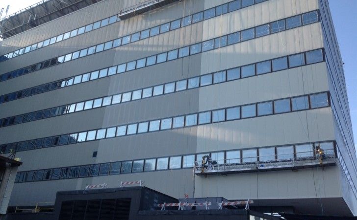 Window cleaners work on a hanging platform attached to the side of a tall, modern office building.